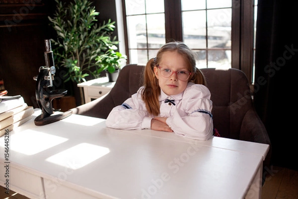 Fototapeta A 7-year-old child in glasses and dressed  school uniform poses at the school table indoor
