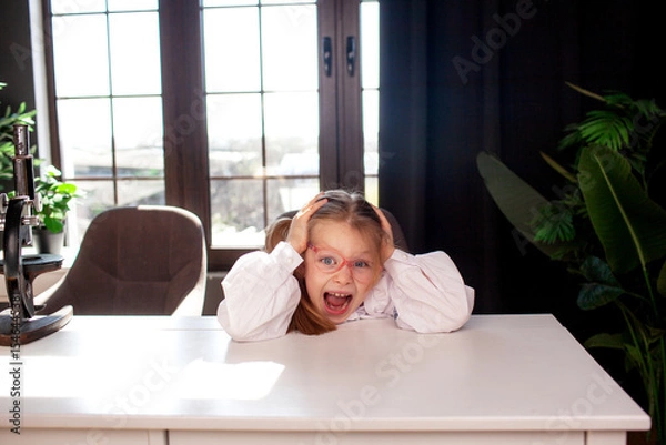 Fototapeta A 7-year-old child in glasses and dressed  school uniform poses at the school table indoor