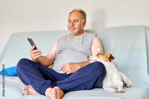 Fototapeta Mature man with diabetes and continuous glucose monitor on his arm using mobile app for checking his health, while sitting in sofa with Jack Russell Terrier dog at home