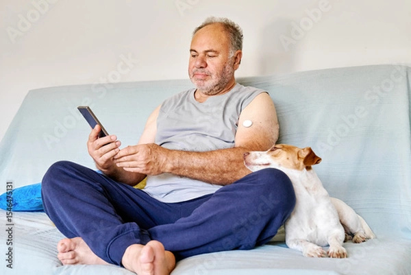 Fototapeta Mature man with diabetes and continuous glucose monitor on his arm using mobile app for checking his health, while sitting in sofa with Jack Russell Terrier dog at home