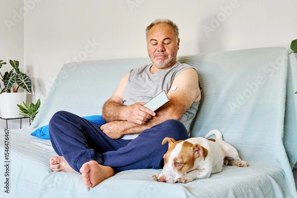 Fototapeta Mature man with diabetes and continuous glucose monitor on his arm using mobile app for checking his health, while sitting in sofa with Jack Russell Terrier dog at home