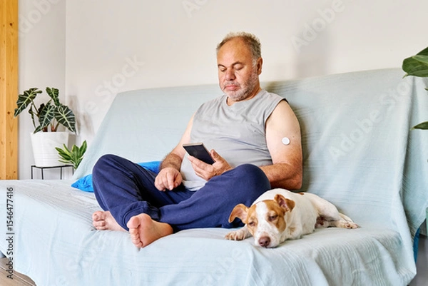 Fototapeta Mature man with diabetes and continuous glucose monitor on his arm using mobile app for checking his health, while sitting in sofa with Jack Russell Terrier dog at home