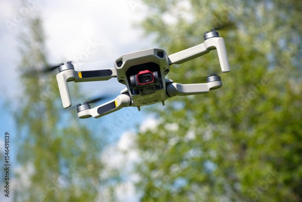 Fototapeta Small Flying Qunmanned aerial vehicle in flight mode with camera outdoor on sunny day summer time blue green garden background