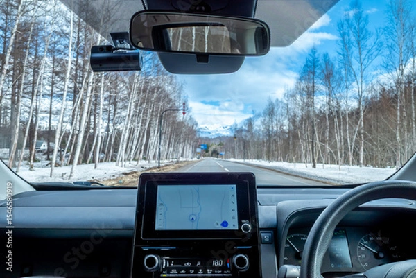 Fototapeta Front view in car with dashboard, navigator on screen and steering wheel, with outside view of leafless trees along the road with snow under cloudy blue sky in winter season