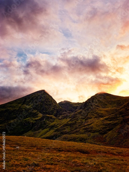 Fototapeta Serene sunset paints the rugged peaks of the Highlands in vibrant hues, casting a warm glow on the tranquil valley below. A solitary sheep grazes peacefully.