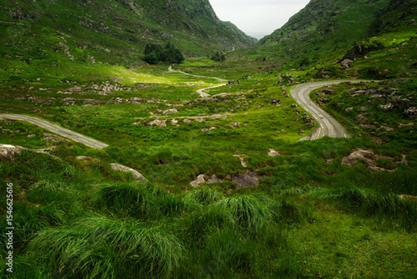 Fototapeta A winding road meanders through rolling hills and towering mountains; a journey awaits.
