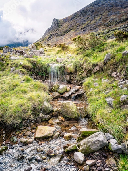 Fototapeta A small waterfall cascades over rocks and vegetation in a picturesque landscape. This is along the popular hiking trail of Carrauntoohill in County Kerry, Ireland, with a mountain in the background.