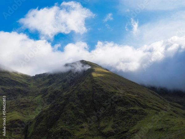 Fototapeta View of rugged Carrauntoohill mountain partially shrouded in clouds against a bright blue sky in County Kerry, Ireland.