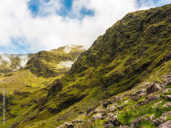 Fototapeta Hills covered in green vegetation and scattered rocks form an Irish mountain landscape. Mist and clouds roll through the valleys near Carrauntoohil along the hiking trail in County Kerry.