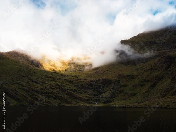 Fototapeta A still body of water is nestled between rolling, grass covered hills that rise into a mountain partially shrouded in white fog near Lough Callee in the Irish countryside.