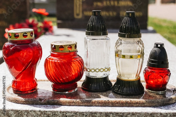 Fototapeta Grave candles with colorful glass. Traditional cemetery lanterns with red and white light. Symbol of memory and prayer. Candles placed on granite gravestone. Honoring the dead. Cemetery decoration.