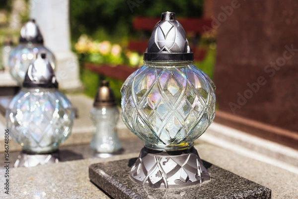 Fototapeta Glass grave candle on tombstone. Shiny lantern with metallic base. Decorative cemetery lamp in sunlight. Memorial light at cemetery. Symbol of memory and respect. Death and remembrance concept.