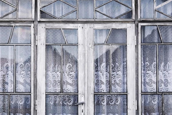 Fototapeta Old wooden door with glass windows. Decorative glass with lace curtains pattern. Vintage entrance white peeling paint. Retro architecture element. Rustic style detail. Traditional house window view.
