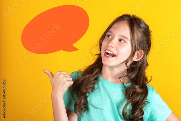 Obraz Portrait of cute young caucasian european blonde girl with opened mouth pointing thumb finger to the side side with empty blank orange speech bubble balloon dialog on yellow background. Studio shot
