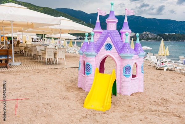 Fototapeta Pink plastic fairy-tale castle on beach sand close-up. Seaside playground with loungers, umbrellas, and mountains in background. Copy space