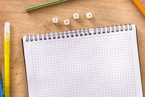 Fototapeta Top view of empty squared notebook with colorful pencils and wooden cubes forming the word idea on desk background