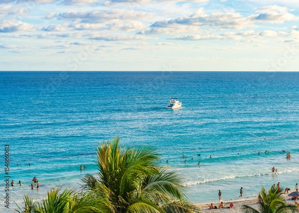 Fototapeta People enjoying the beach and swimming in the clear blue waters of Cancun Hotel Zone with a boat cruising offshore under a partly cloudy sky High-quality photo.