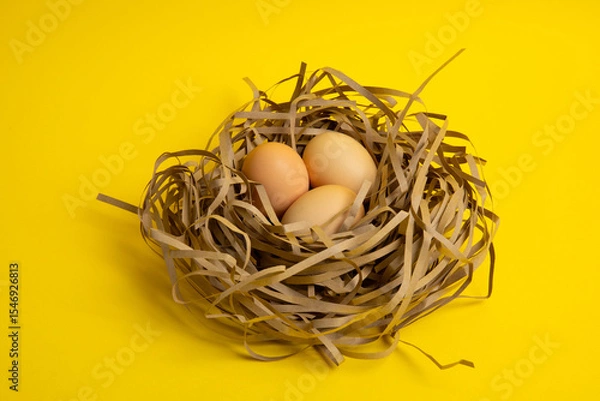 Obraz Three brown eggs in paper nest – yellow background, top view