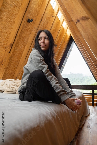 Fototapeta A young woman sits peacefully cross-legged in a lotus pose on a cozy bed inside a rustic glamping tent, bathed in soft natural light, embracing calm and mindfulness.

