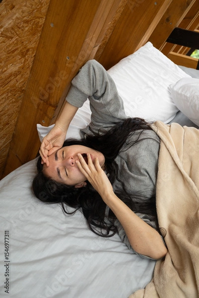 Fototapeta A young woman rises from bed, yawning and stretching with her hand on her forehead, bathed in soft morning light inside a warm and rustic glamping tent.

