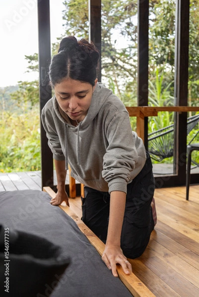 Fototapeta A young woman kneels beside a wooden bed in a cozy cabin with large glass windows. The scene evokes calmness, order, and a deep connection with nature.
