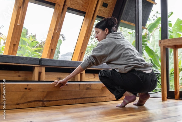 Fototapeta Seen from behind, a young woman squats as she opens a drawer beneath a wooden bed in a cozy cabin. The scene captures a quiet domestic moment, emphasizing order, presence, and harmony with the natural