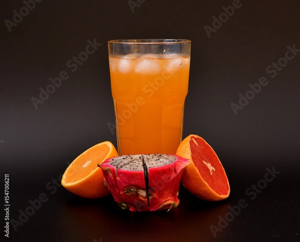 Obraz Tropical fruit juice in a tall glass with ice on a black background, next to it are pieces of ripe orange, grapefruit and pitaya.