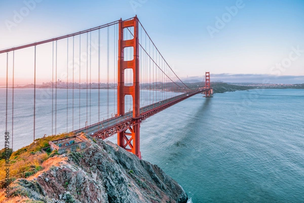 Fototapeta Golden Gate Bridge at sunrise in San Francisco, California. Scenic view from Battery Spencer with soft pastel sky, calm bay waters, and iconic red architecture. Tranquil travel landscape.