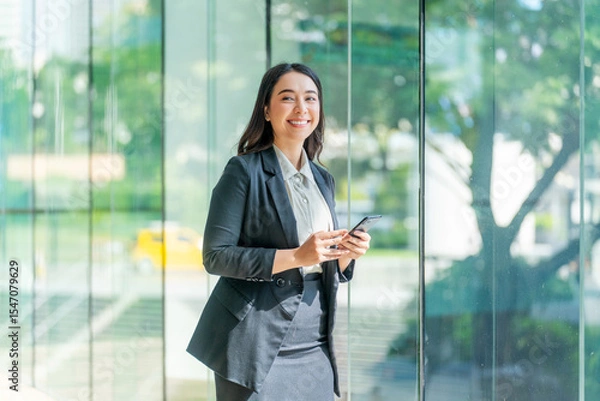 Obraz Asian businesswoman using smartphone near glass wall with reflection. Confident woman in formal outfit using phone in modern indoor space.