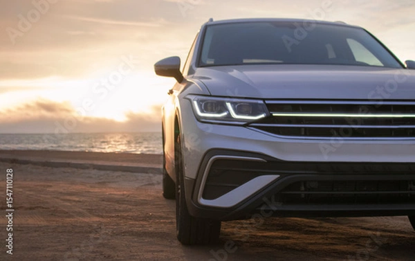 Fototapeta Silhouetted by a colorful sunset, a gray car stands near the ocean's edge. The warm light and open space capture a moment of solitude and exploration. 