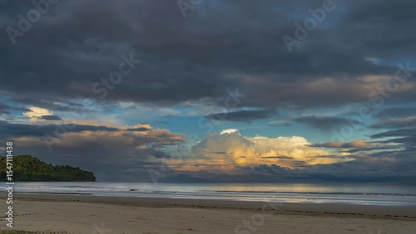 Fototapeta Sunset on a tropical island. The clouds in the blue evening sky are highlighted in gold and pink. Ocean waves are spreading across the sandy beach. Reflection on the sea surface.A hill in the distance
