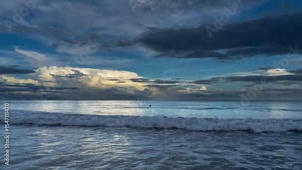 Fototapeta Early morning by the ocean. The waves roll towards the shore, foam and spread. A swimming man's head is visible in the distance. The clouds in the blue sky are illuminated with golden light.Reflection