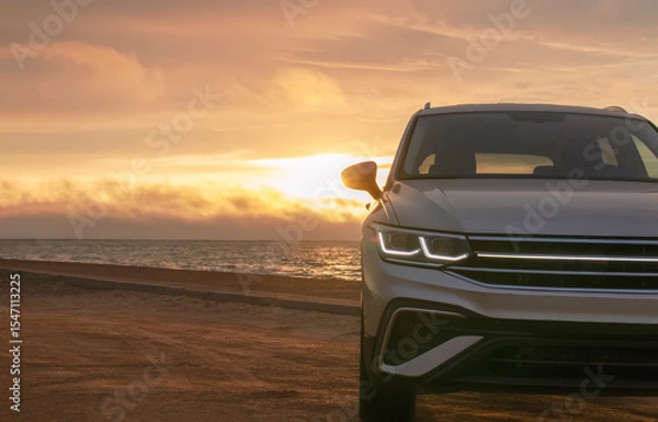 Fototapeta Silhouetted by a colorful sunset, a gray car stands near the ocean's edge. The warm light and open space capture a moment of solitude and exploration. 
