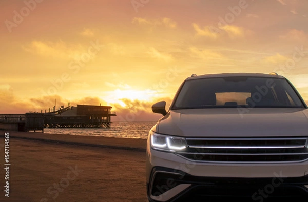 Fototapeta Silhouetted by a colorful sunset, a gray car stands near the ocean's edge. The warm light and open space capture a moment of solitude and exploration. 