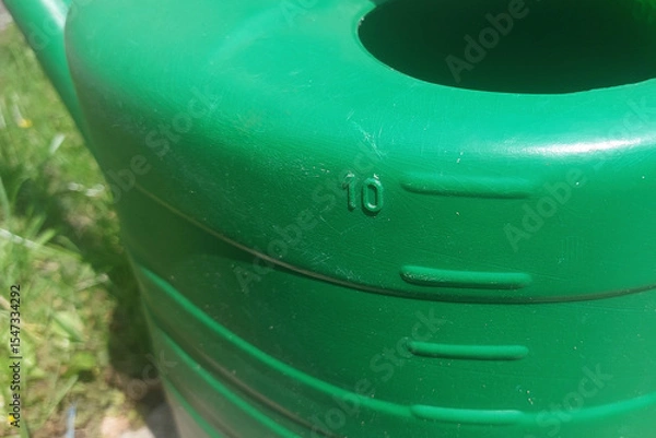 Fototapeta Green watering can with a capacity of 10 liters placed on a sunny garden path surrounded by grass