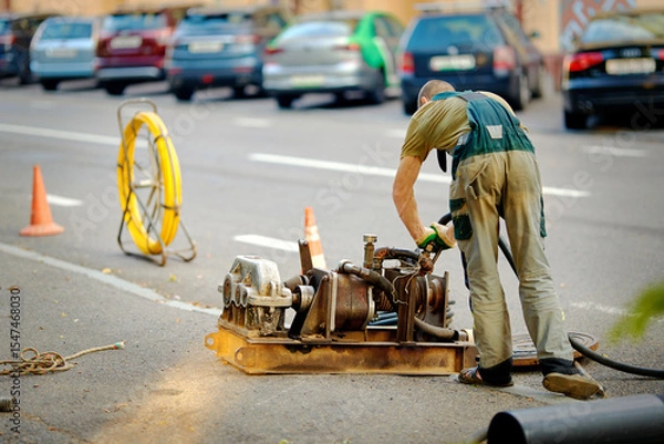 Fototapeta Utility worker using cable pulling rod tool to guide fiber optic cables through an open manhole during sewer system installation and underground network repair. Fiber optic installation in progress