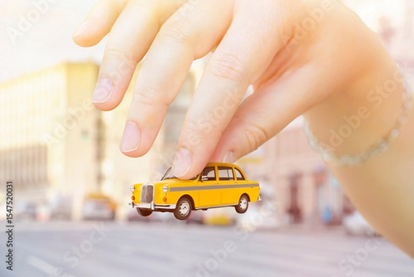 Fototapeta Close-up of a hand holding a toy taxi car. Background on the topic of taxi service cost and amenities.