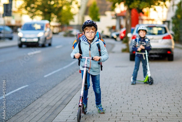 Fototapeta Two school kid boys in safety helmet riding with scooter in the city with backpack on sunny day. Happy children in colorful clothes biking on way to school.