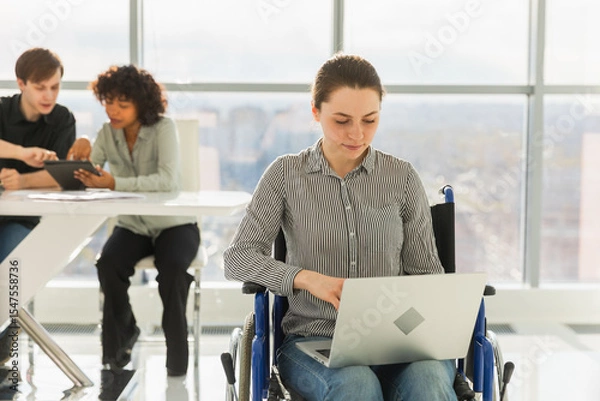 Fototapeta Accessible environment. Woman in chair for people with disability working with laptop in modern office. Diverse multiracial group of people colleagues working together. Young professional team