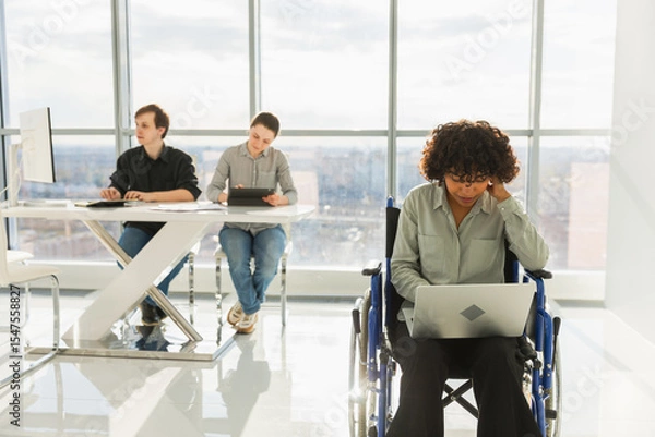 Fototapeta Accessible environment. African woman in chair for people with disability working with laptop in modern office. Diverse multiracial group of people colleagues working together. Young professional team