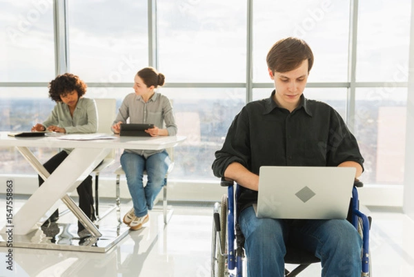 Fototapeta Accessible environment. Young man in chair for people with disability working with laptop in modern office. Diverse multiracial group of people colleagues working together. Young professional team