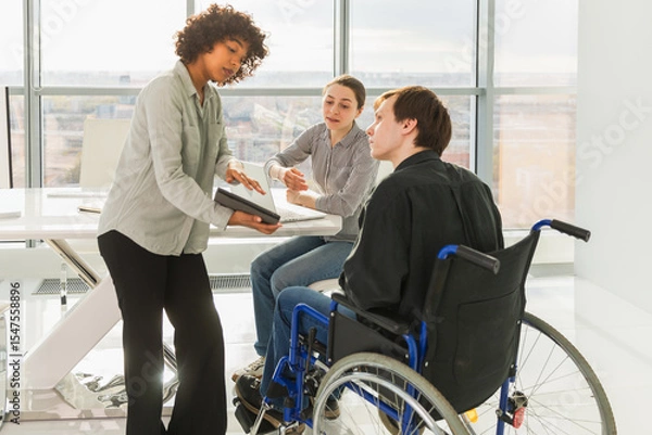 Fototapeta Teamwork brainstorm. Diverse multiracial colleagues man in chair for people with disability working together. Group of people sitting in creative office discussing business. Young professional team