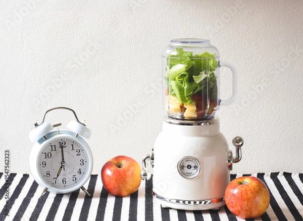 Fototapeta white vintage blender or smoothie maker machine  with vegetables and fruit inside, white vintage alarm clock shows 7 o'clock and 2 apples  on  black and white stripe table cloth and white wall.