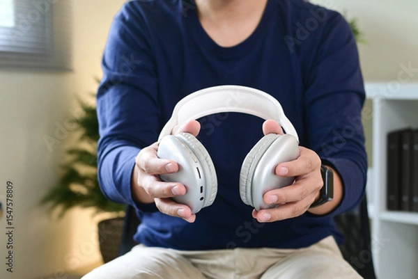Obraz A man is holding a pair of headphones in his hands. The man is sitting on a chair, and there are several books around him