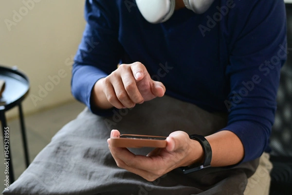 Obraz A man is sitting on a couch, holding a cell phone in his hands and looking at it intently. He is focused on the screen, possibly checking his messages or browsing the internet.