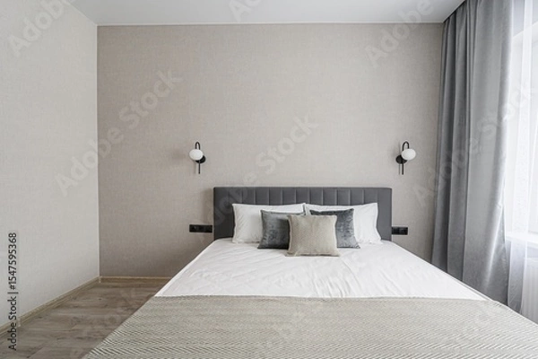 Fototapeta serene bedroom with a gray headboard bed, white linens, and neutral-toned walls. Symmetrical wall lamps frame the bed. Curtains filter light