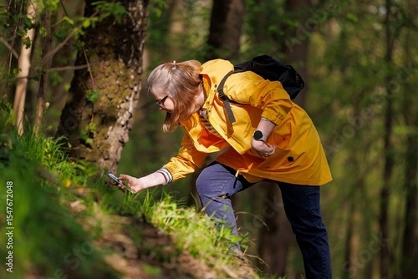 Fototapeta A person in a bright yellow jacket examines the ground intently while surrounded by lush green foliage.
