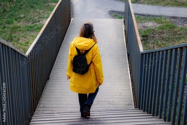 Fototapeta A person wearing a vibrant yellow raincoat walks down wooden steps, surrounded by lush greenery in a serene park.