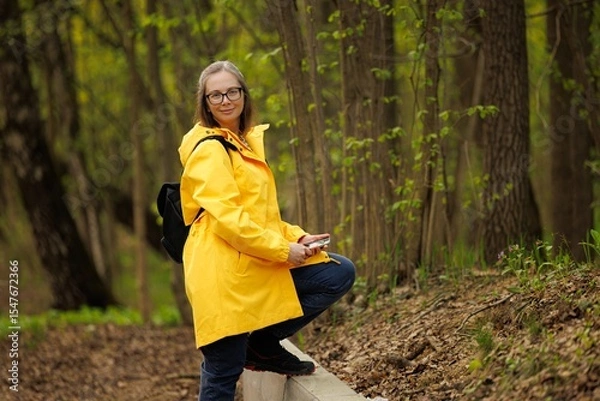 Fototapeta A woman happily poses on a trail, dressed in a yellow rain jacket, surrounded by fresh greenery and trees.