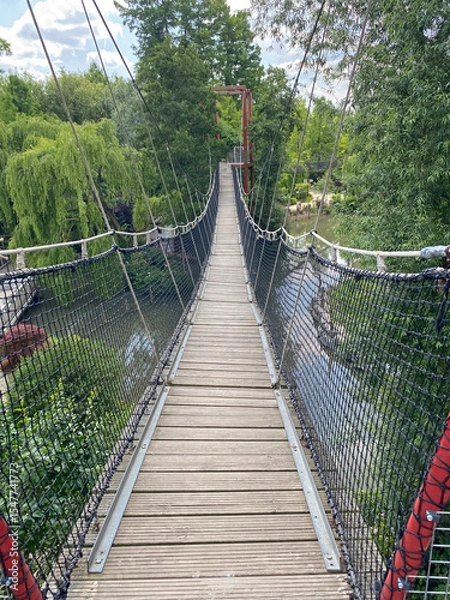 Obraz Perspective view of a suspension rope bridge (monkey bridge) entrance crossing over a river, highlighting the structure’s length and natural surroundings